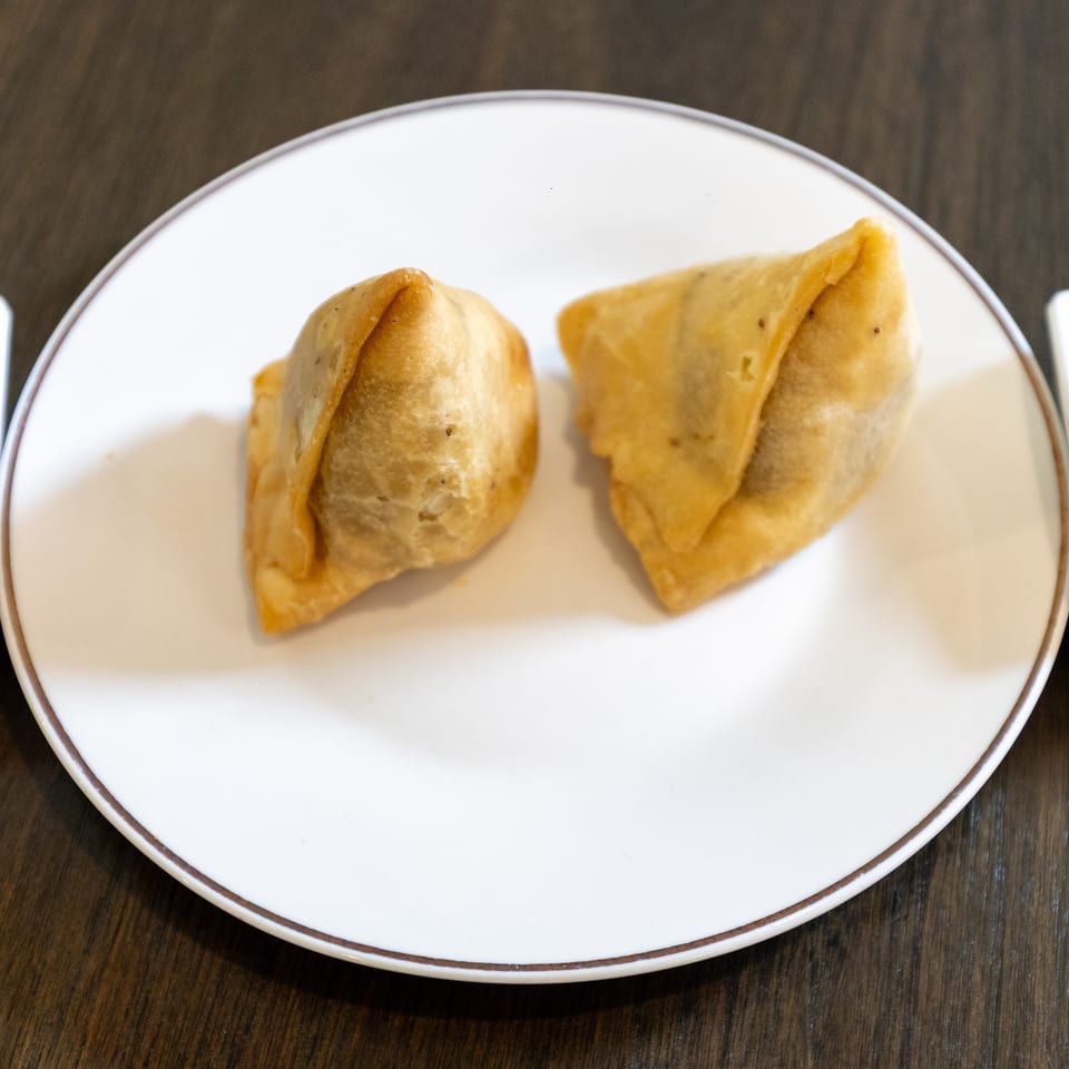 Two golden fried samosas on a white plate, served with green and brown dipping sauces on a wooden table.
