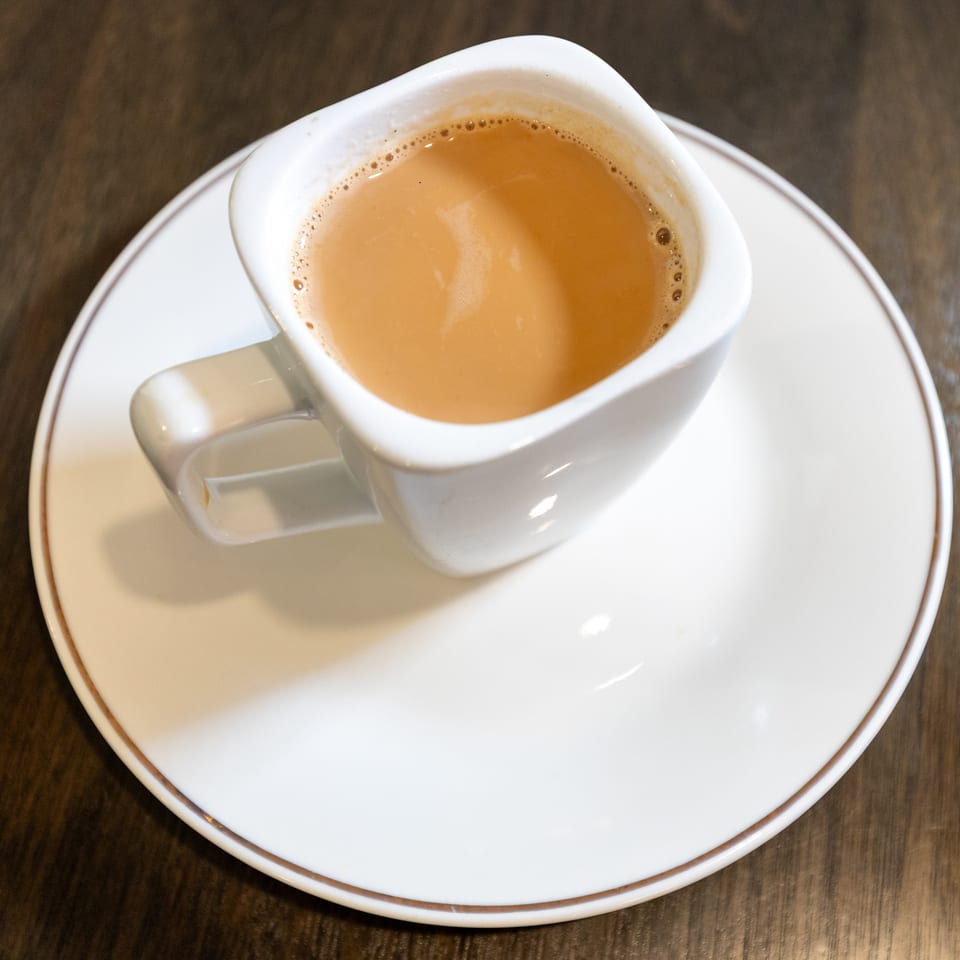 White square cup filled with frothy coffee sits on a matching saucer atop a dark wooden table.