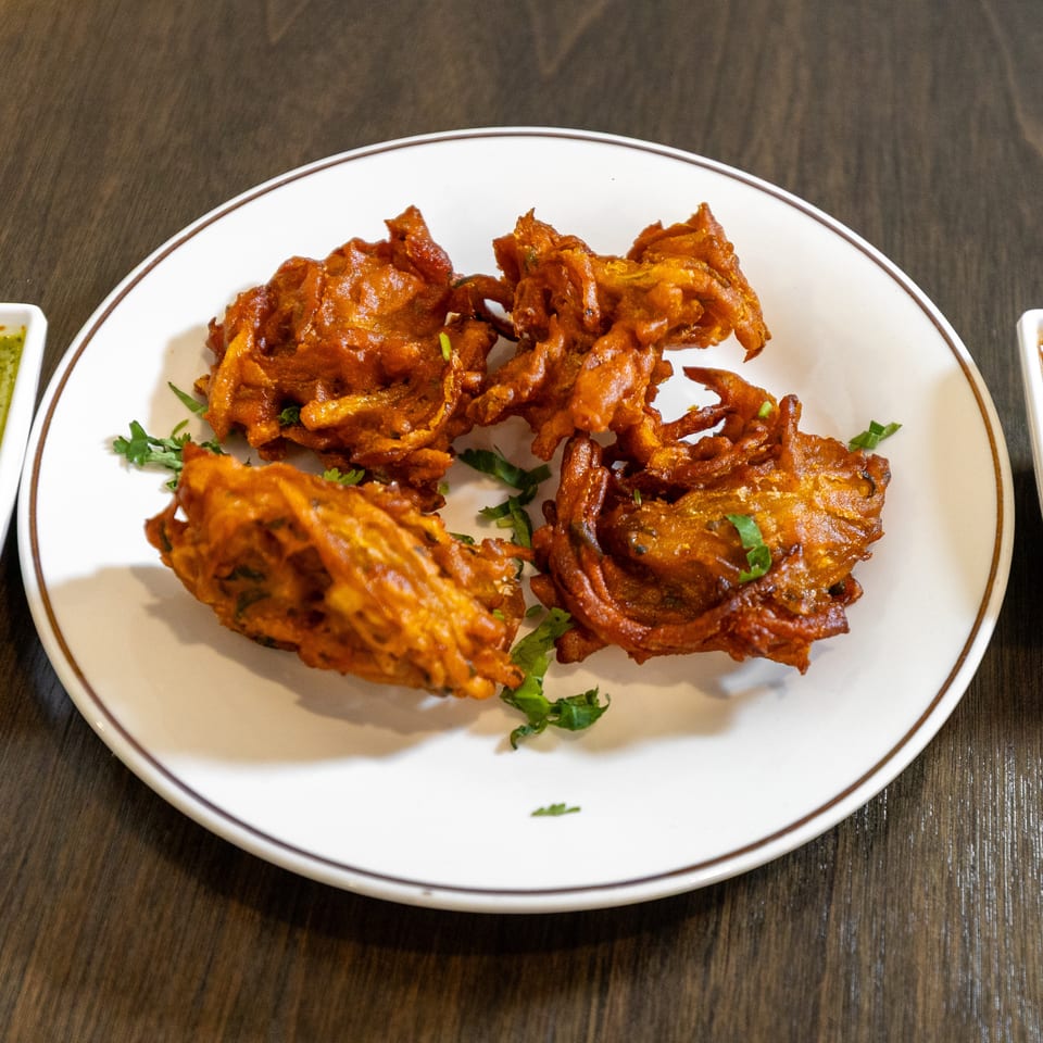 Crispy golden vegetable fritters garnished with cilantro on a white plate, served with green chutney and brown dipping sauce.