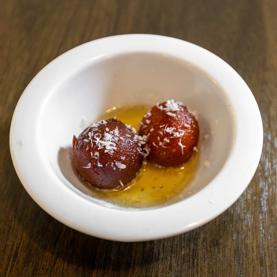 Three syrup-soaked brown dessert balls topped with shredded coconut sit in a white bowl on a wooden table.