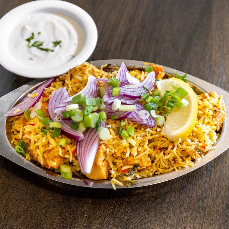 Spiced chicken biryani topped with red onion, scallions, and lemon, served with creamy raita on a metal platter.