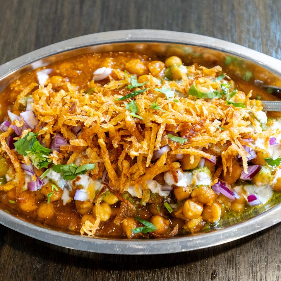 Indian chaat topped with chickpeas, yogurt, chutneys, crispy fried onions, red onion, and cilantro in a metal platter.