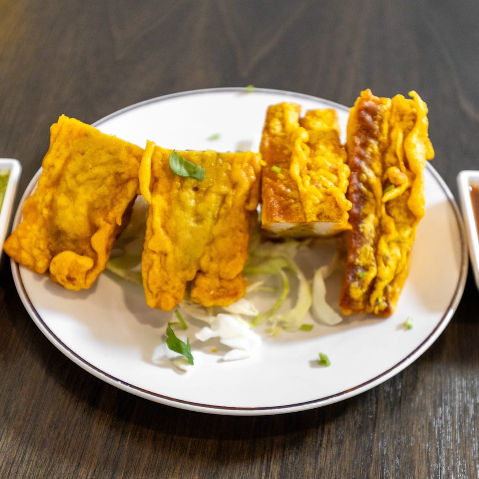 Golden-fried Indian appetizers arranged on a white plate with green chutney, brown sauce, cabbage, and cilantro garnish.