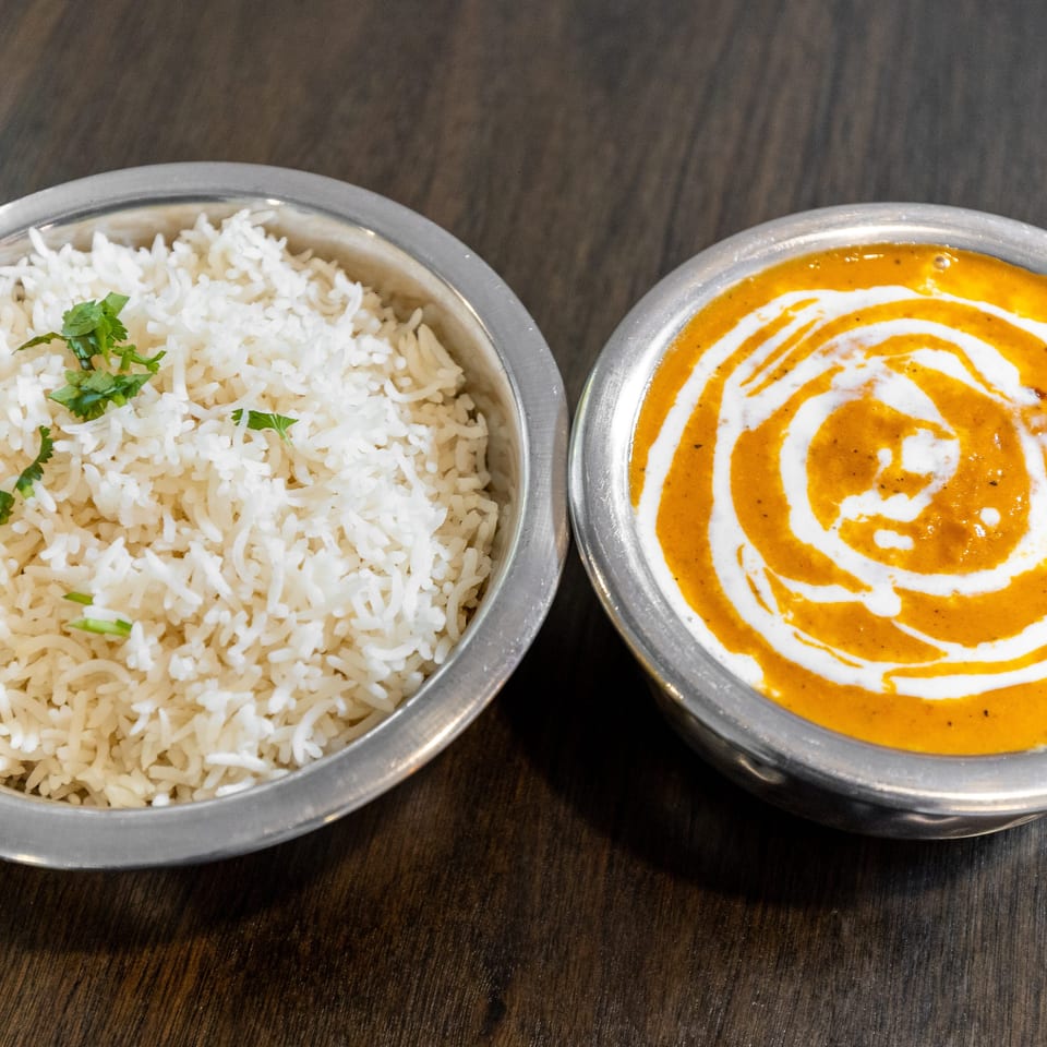 Steamed white rice garnished with cilantro beside creamy orange curry drizzled with white cream in metal bowls.
