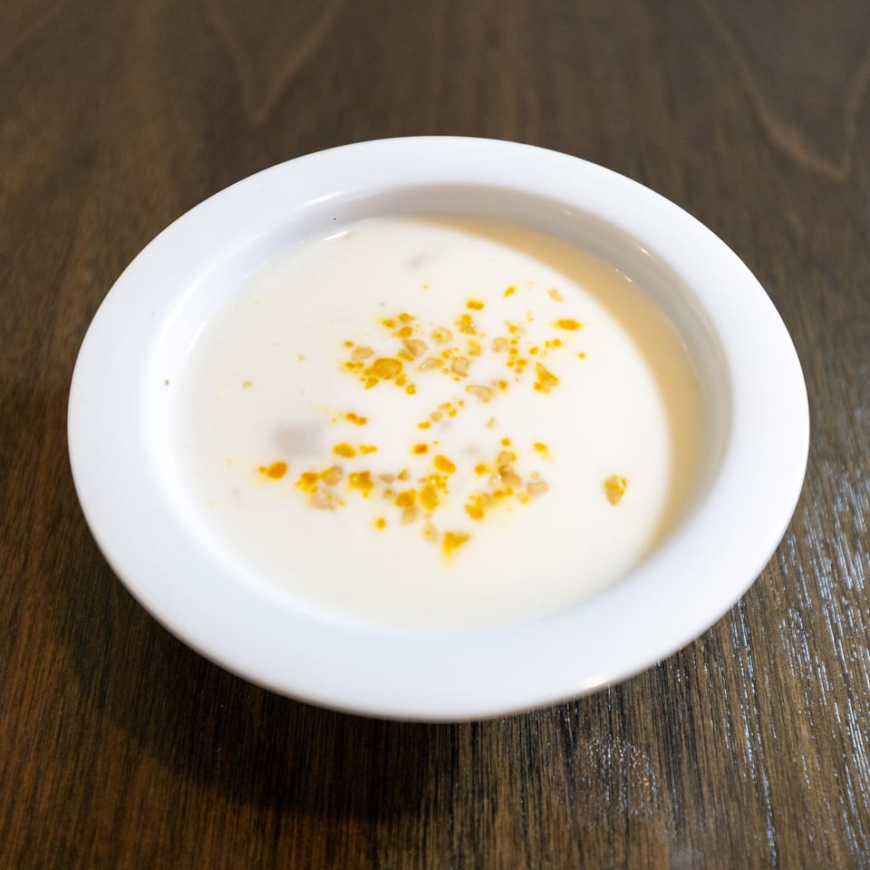 Creamy white soup topped with golden oil droplets and spices in a white bowl on a wooden table.